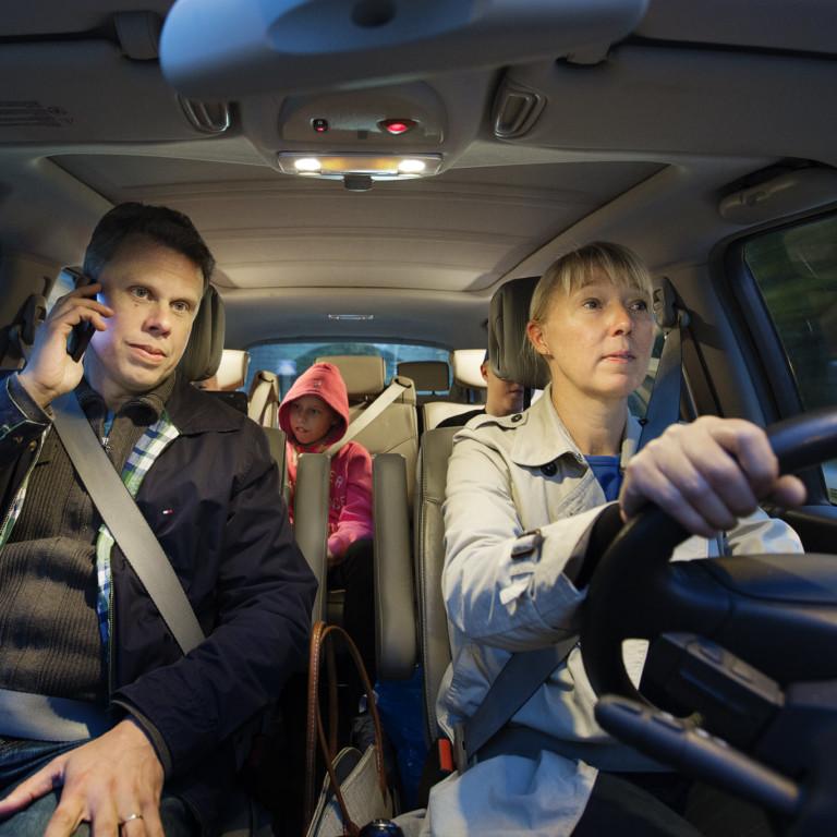 Kuvassa apukuljettaja autossa puhuu puhelimeen kuljettajan keskittyessä ajamiseen.