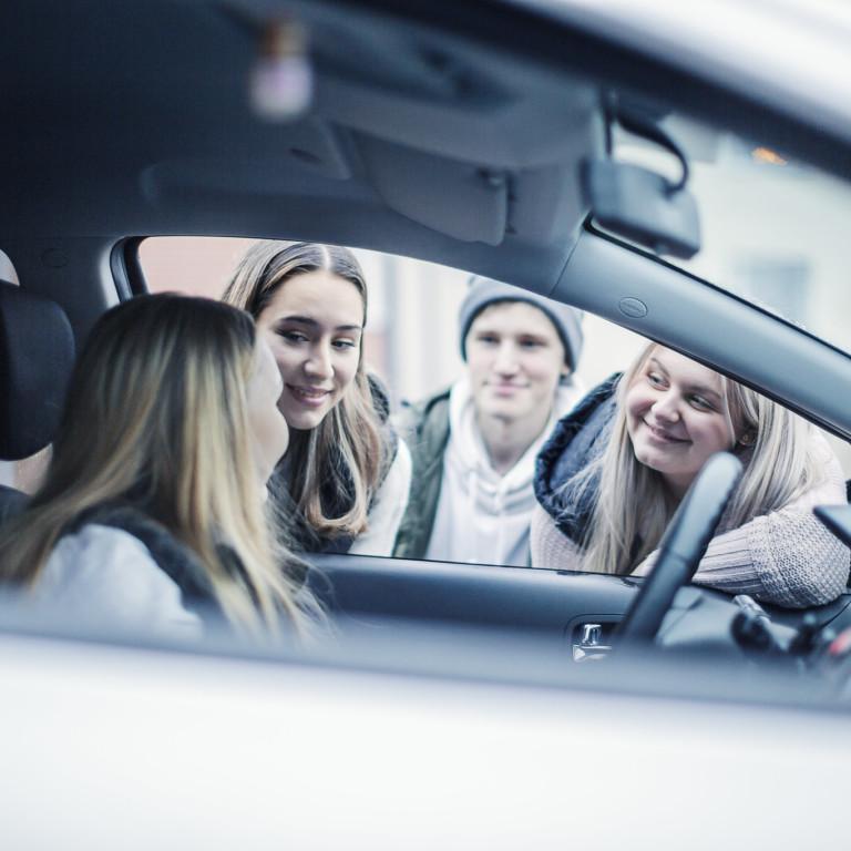 Kuvassa nuori kuljettaja pysähtyneessä henkilöautossa, muita nuoria juttelee hänelle ikkunan kautta.